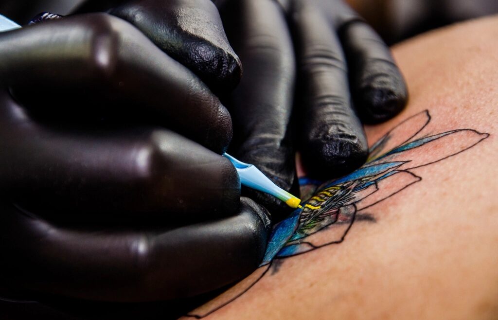 Black-gloved hands filling a tattoo with yellow and blue ink
